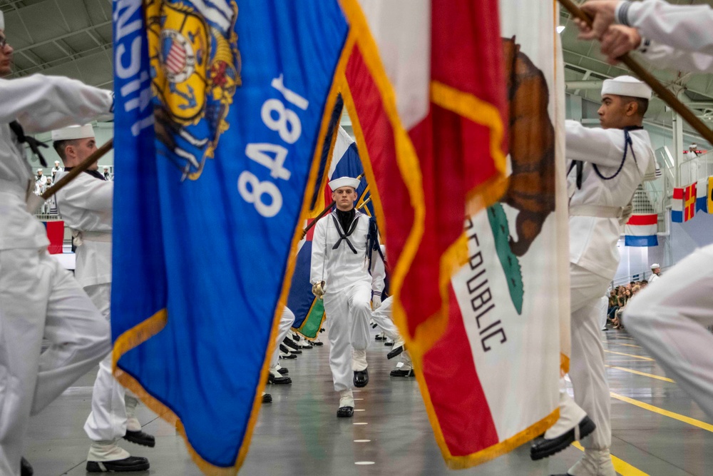 U.S. Navy sailors in dress white uniforms are participating in a formal color guard or graduation ceremony inside a large indoor facility. Multiple state flags are prominently displayed, including the California Republic flag and what appears to be the Wisconsin state flag, along with a U.S. Navy flag dated 1848. Sailors are marching in formation while carrying flags on staffs in a ceremonial drill.
