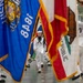 U.S. Navy sailors in dress white uniforms are participating in a formal color guard or graduation ceremony inside a large indoor facility. Multiple state flags are prominently displayed, including the California Republic flag and what appears to be the Wisconsin state flag, along with a U.S. Navy flag dated 1848. Sailors are marching in formation while carrying flags on staffs in a ceremonial drill.