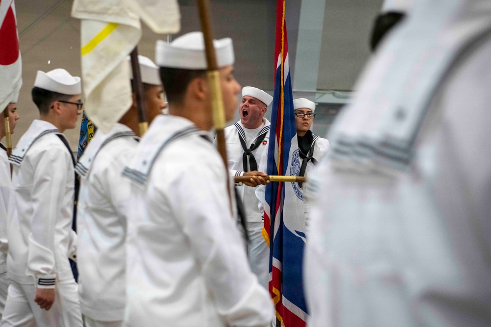 A military naval ceremony or parade is taking place indoors, featuring personnel dressed in white Navy uniforms with black neckerchiefs and white sailor caps. A color guard is present, carrying flags including what appears to be a British Union Jack-style flag. One individual in the center appears to be calling out commands or cadence with mouth open wide.