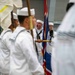 A military naval ceremony or parade is taking place indoors, featuring personnel dressed in white Navy uniforms with black neckerchiefs and white sailor caps. A color guard is present, carrying flags including what appears to be a British Union Jack-style flag. One individual in the center appears to be calling out commands or cadence with mouth open wide.