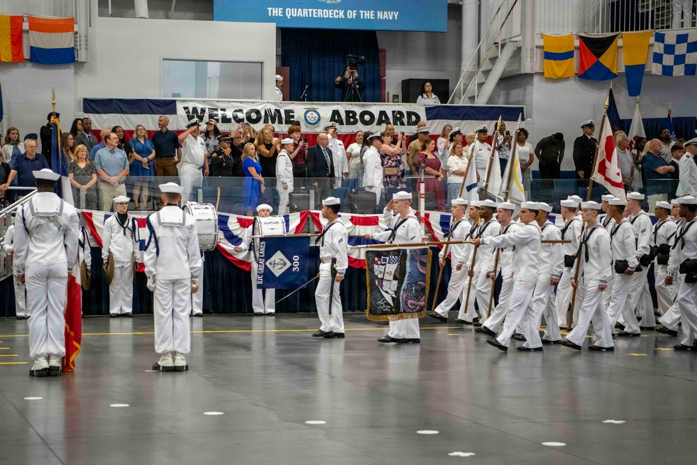 A U.S. Navy graduation or passing-out ceremony is taking place inside an indoor drill hall identified as 'The Quarterdeck of the Navy.' Navy recruits dressed in white dress uniforms are marching in formation while carrying flags and banners, including one marked 'RTC Great Lakes 300.' Spectators and officials observe from a raised gallery area beneath a large 'Welcome Aboard' banner.