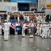 A U.S. Navy graduation or passing-out ceremony is taking place inside an indoor drill hall identified as 'The Quarterdeck of the Navy.' Navy recruits dressed in white dress uniforms are marching in formation while carrying flags and banners, including one marked 'RTC Great Lakes 300.' Spectators and officials observe from a raised gallery area beneath a large 'Welcome Aboard' banner.