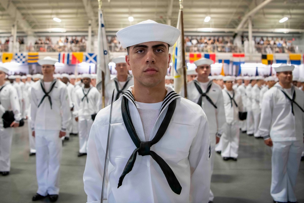 A formal U.S. Navy recruit graduation ceremony taking place inside a large indoor facility. Multiple rows of recruits in white dress uniforms stand at attention in formation, with the foreground recruit serving as a guidon bearer holding a ceremonial flag. Spectators are visible in elevated bleachers in the background, and naval signal flags decorate the venue.