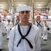 A formal U.S. Navy recruit graduation ceremony taking place inside a large indoor facility. Multiple rows of recruits in white dress uniforms stand at attention in formation, with the foreground recruit serving as a guidon bearer holding a ceremonial flag. Spectators are visible in elevated bleachers in the background, and naval signal flags decorate the venue.