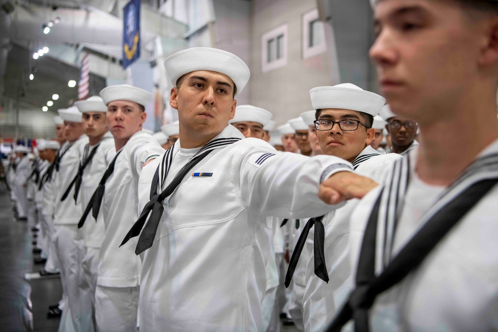 A formation of U.S. Navy sailors in dress white uniforms are standing at attention or in a marching formation inside a large indoor facility. The sailors are wearing traditional white Crackerjack uniforms with black neckerchiefs and white Dixie cup hats. The setting appears to be a military graduation or pass-in-review ceremony.