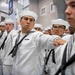A formation of U.S. Navy sailors in dress white uniforms are standing at attention or in a marching formation inside a large indoor facility. The sailors are wearing traditional white Crackerjack uniforms with black neckerchiefs and white Dixie cup hats. The setting appears to be a military graduation or pass-in-review ceremony.