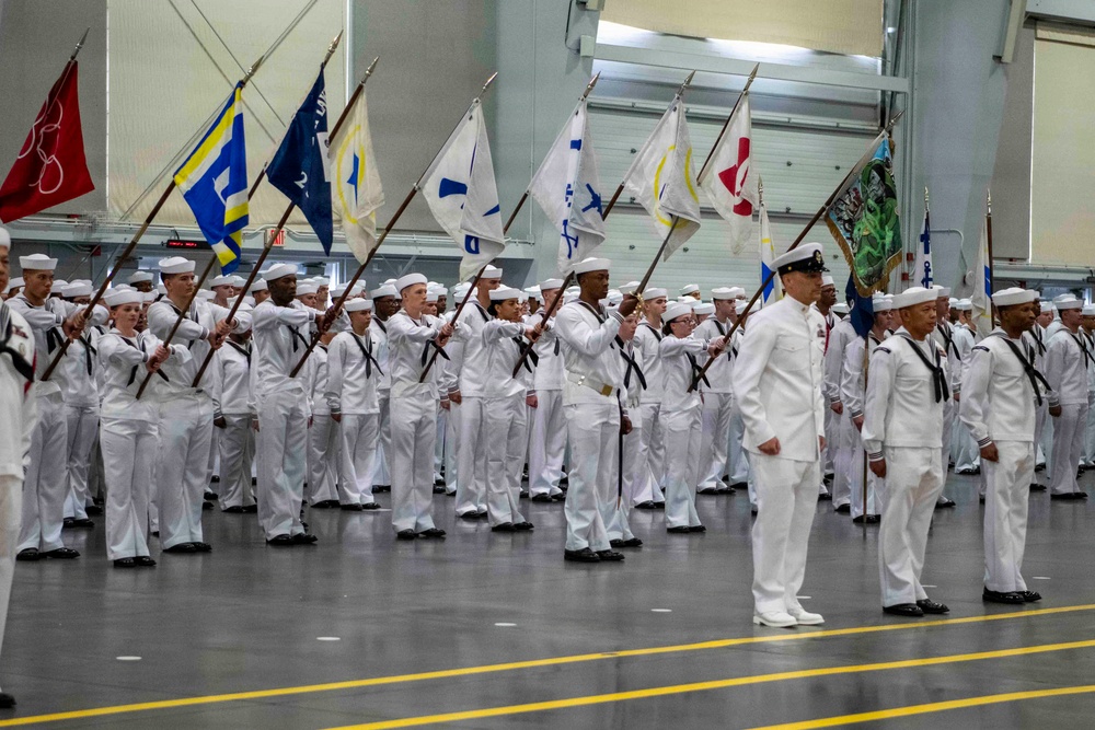 A large formation of U.S. Navy sailors dressed in white dress uniforms are participating in an indoor military ceremony, likely a graduation or pass-in-review event. Multiple sailors in the formation are carrying unit guidon flags on long poles, displaying various colors and insignia. Two senior personnel stand at attention in the foreground, appearing to oversee or lead the formation.