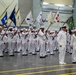 A large formation of U.S. Navy sailors dressed in white dress uniforms are participating in an indoor military ceremony, likely a graduation or pass-in-review event. Multiple sailors in the formation are carrying unit guidon flags on long poles, displaying various colors and insignia. Two senior personnel stand at attention in the foreground, appearing to oversee or lead the formation.