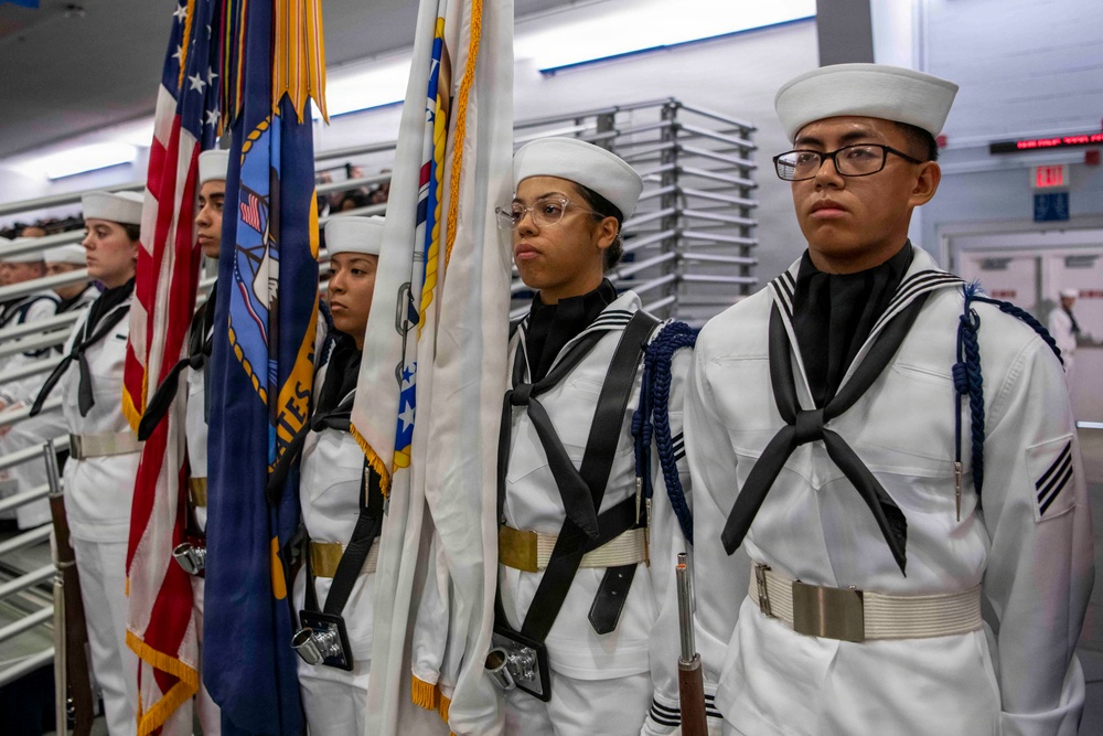 A U.S. Navy color guard stands at attention in dress white uniforms during a formal military ceremony, likely at a naval training facility. The personnel are carrying ceremonial flags including the American flag and what appears to be a U.S. Navy flag. The setting appears to be an indoor facility, possibly a mess hall or large ceremonial room.