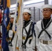 A U.S. Navy color guard stands at attention in dress white uniforms during a formal military ceremony, likely at a naval training facility. The personnel are carrying ceremonial flags including the American flag and what appears to be a U.S. Navy flag. The setting appears to be an indoor facility, possibly a mess hall or large ceremonial room.