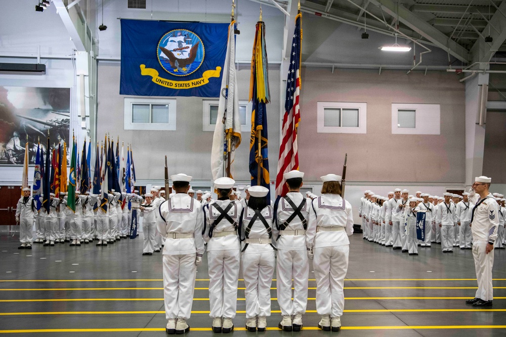 A formal United States Navy graduation or pass-in-review ceremony taking place inside a large indoor drill hall. Multiple sailors in dress white uniforms are assembled in formation, with a color guard bearing flags at the center. A large United States Navy flag with the official seal is prominently displayed on the back wall.