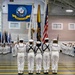 A formal United States Navy graduation or pass-in-review ceremony taking place inside a large indoor drill hall. Multiple sailors in dress white uniforms are assembled in formation, with a color guard bearing flags at the center. A large United States Navy flag with the official seal is prominently displayed on the back wall.