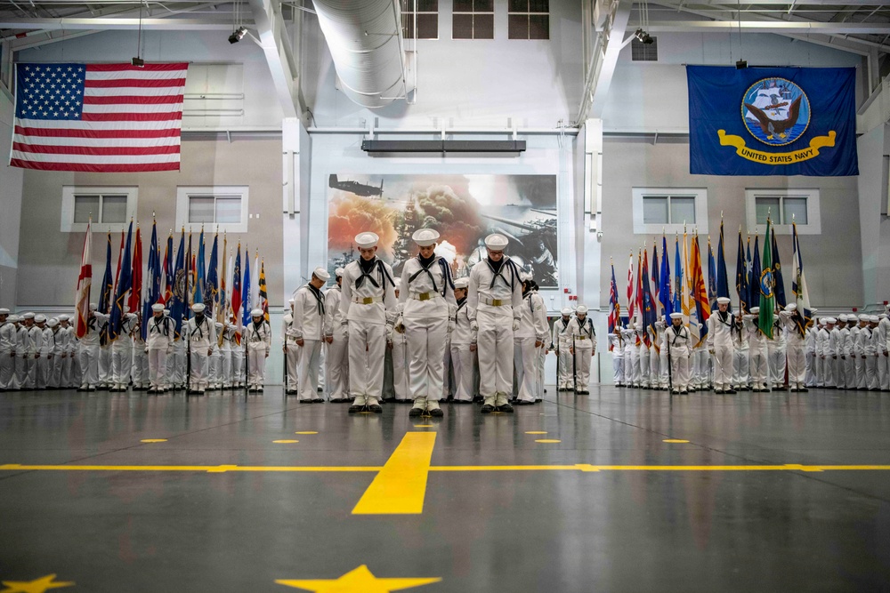 A formal United States Navy graduation or pass-in-review ceremony taking place inside a large indoor drill hall. Navy recruits dressed in white dress uniforms are arranged in formation, with a small group of sailors bowing or receiving recognition at the center. The hall is decorated with the American flag, the United States Navy flag, and numerous state and military flags along both sides.