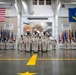 A formal United States Navy graduation or pass-in-review ceremony taking place inside a large indoor drill hall. Navy recruits dressed in white dress uniforms are arranged in formation, with a small group of sailors bowing or receiving recognition at the center. The hall is decorated with the American flag, the United States Navy flag, and numerous state and military flags along both sides.