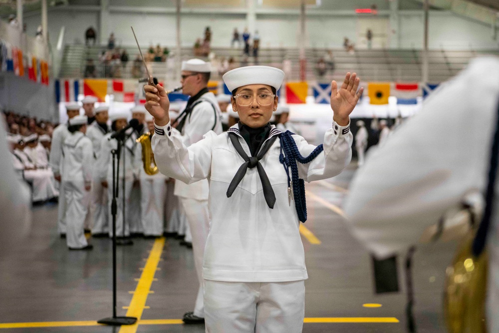 A U.S. Navy band conductor in dress white uniform leads a musical ensemble during what appears to be a formal military ceremony or graduation event held in a large indoor facility. The conductor, wearing glasses and a blue aiguillette, holds a baton in one hand while gesturing with the other. Behind them, rows of sailors in white uniforms and band members with instruments are visible, with colorful signal flags decorating the background.