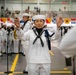 A U.S. Navy band conductor in dress white uniform leads a musical ensemble during what appears to be a formal military ceremony or graduation event held in a large indoor facility. The conductor, wearing glasses and a blue aiguillette, holds a baton in one hand while gesturing with the other. Behind them, rows of sailors in white uniforms and band members with instruments are visible, with colorful signal flags decorating the background.
