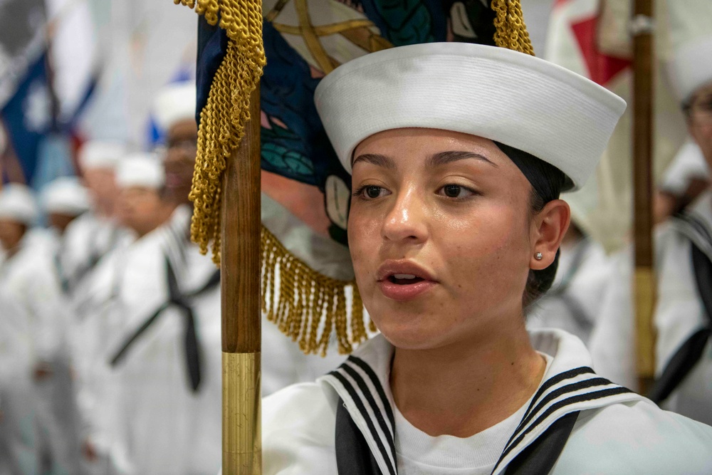 A female sailor in U.S. Navy dress white uniform is holding a ceremonial flag/guidon with gold fringe during what appears to be a formal military ceremony or parade. She is wearing the distinctive white dixie cup cover and a traditional sailor collar with black stripes. Additional sailors in matching dress whites are visible in the blurred background.