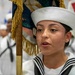 A female sailor in U.S. Navy dress white uniform is holding a ceremonial flag/guidon with gold fringe during what appears to be a formal military ceremony or parade. She is wearing the distinctive white dixie cup cover and a traditional sailor collar with black stripes. Additional sailors in matching dress whites are visible in the blurred background.