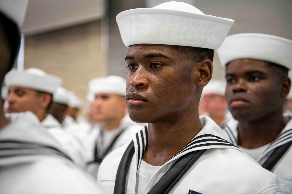 The image shows a group of naval recruits or sailors in dress white uniforms standing in formation. The foreground features a young male sailor in sharp focus wearing the distinctive white Dixie cup cap and traditional navy jumper with black neckerchief. The setting appears to be an indoor ceremony or formal military gathering.