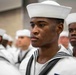 The image shows a group of naval recruits or sailors in dress white uniforms standing in formation. The foreground features a young male sailor in sharp focus wearing the distinctive white Dixie cup cap and traditional navy jumper with black neckerchief. The setting appears to be an indoor ceremony or formal military gathering.