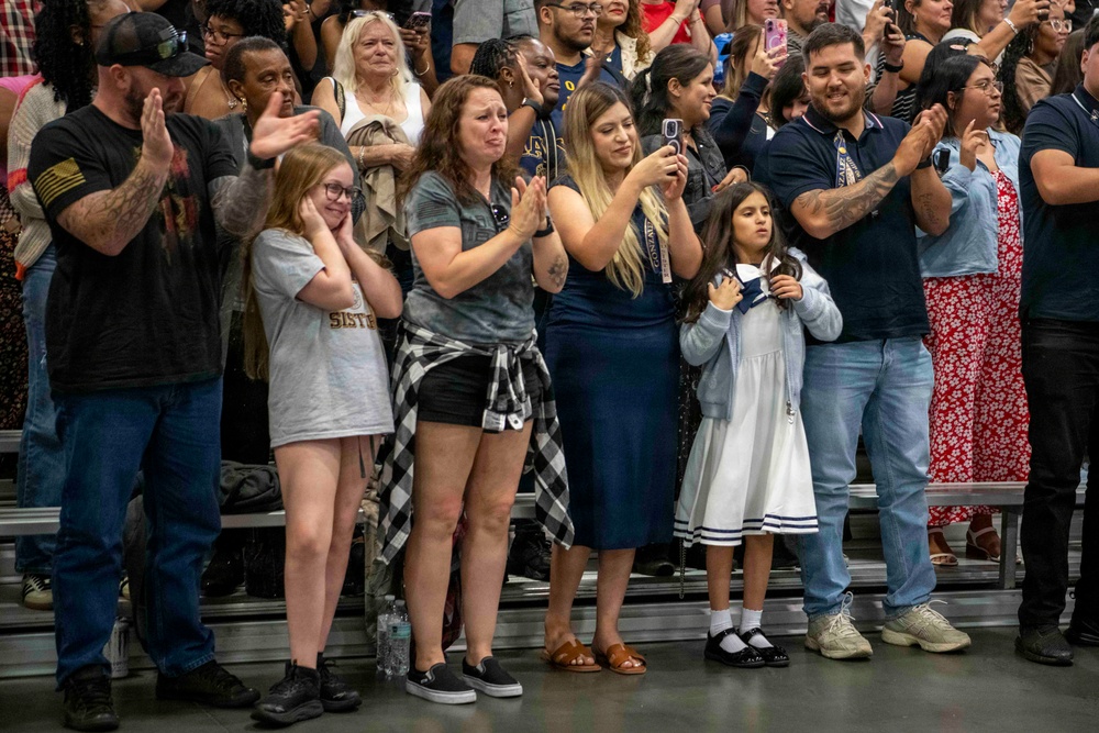 A crowd of people standing on bleachers at what appears to be a military or graduation ceremony, displaying strong emotional reactions including clapping, crying, and taking photos. The audience includes adults and children of various ages, with several individuals visibly moved to tears. The atmosphere suggests a significant and emotionally charged event such as a military homecoming or graduation.