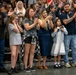 A crowd of people standing on bleachers at what appears to be a military or graduation ceremony, displaying strong emotional reactions including clapping, crying, and taking photos. The audience includes adults and children of various ages, with several individuals visibly moved to tears. The atmosphere suggests a significant and emotionally charged event such as a military homecoming or graduation.