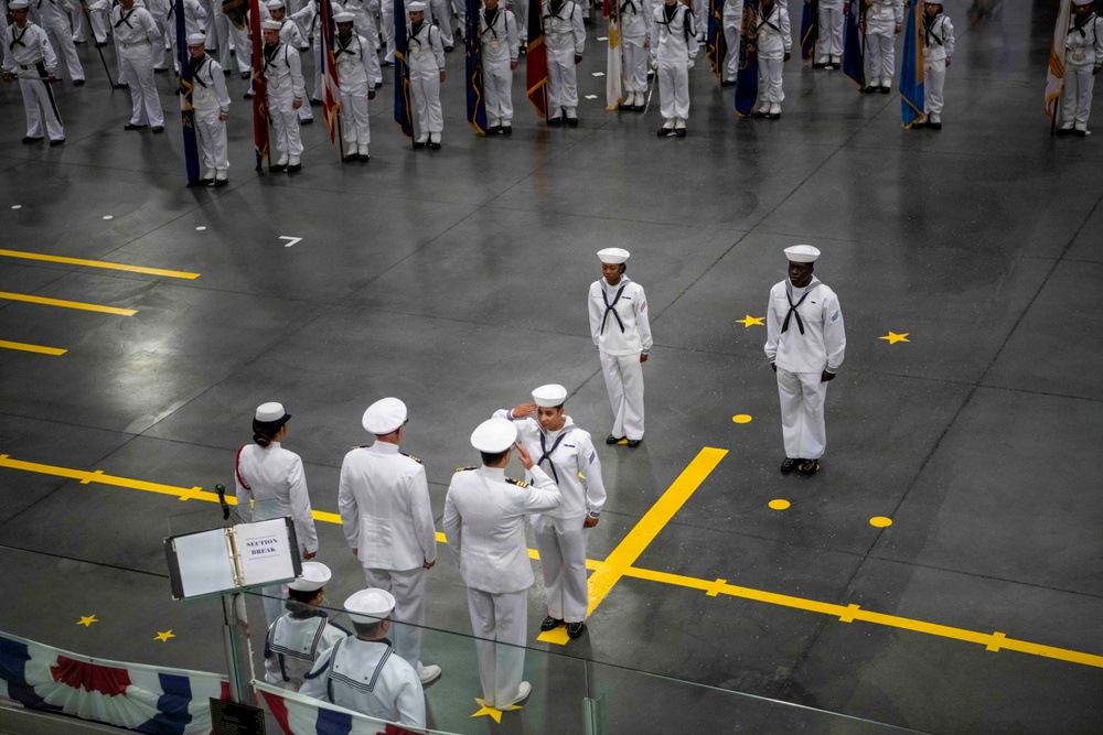A formal U.S. Navy military ceremony is taking place in a large indoor drill hall or hangar facility. Personnel in white dress uniforms are arranged in formation, with color guard units holding flags and an officer receiving a salute in the foreground. The scene appears to be a graduation or pass-in-review ceremony, likely at a Navy recruit training command.