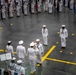 A formal U.S. Navy military ceremony is taking place in a large indoor drill hall or hangar facility. Personnel in white dress uniforms are arranged in formation, with color guard units holding flags and an officer receiving a salute in the foreground. The scene appears to be a graduation or pass-in-review ceremony, likely at a Navy recruit training command.