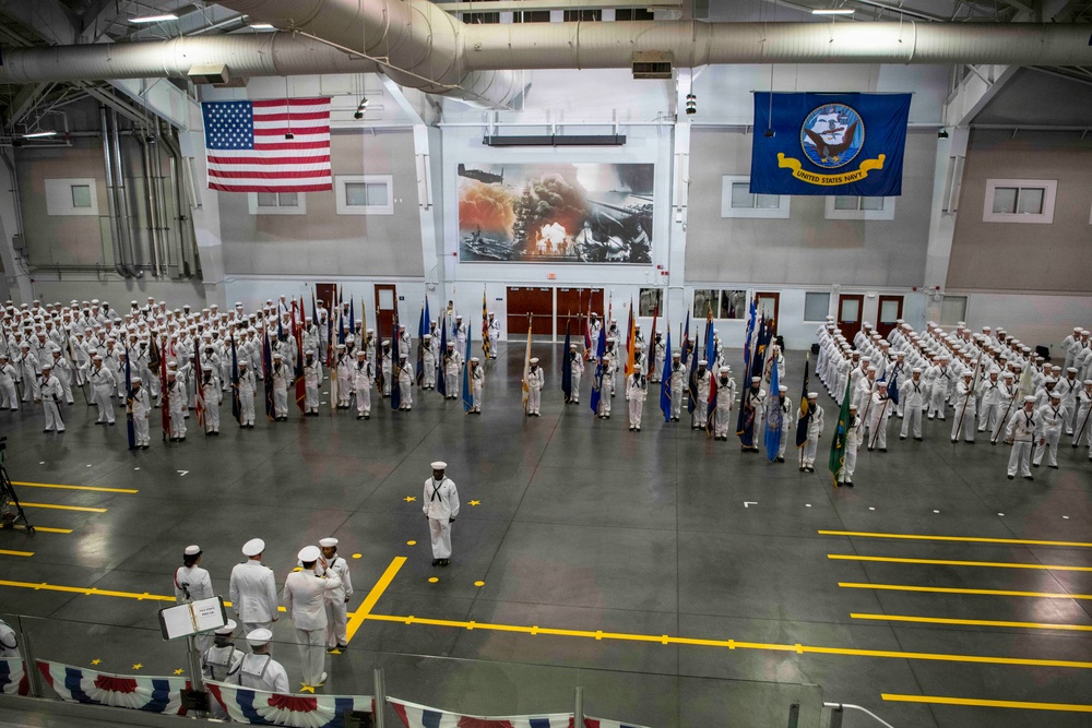 A formal United States Navy military ceremony is taking place inside a large indoor drill hall or hangar facility. Hundreds of Navy personnel dressed in white dress uniforms are assembled in formation, with color guard units holding numerous flags and standards. The venue is decorated with an American flag, a United States Navy flag, and a large mural depicting what appears to be a naval battle scene.