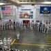 A formal United States Navy military ceremony is taking place inside a large indoor drill hall or hangar facility. Hundreds of Navy personnel dressed in white dress uniforms are assembled in formation, with color guard units holding numerous flags and standards. The venue is decorated with an American flag, a United States Navy flag, and a large mural depicting what appears to be a naval battle scene.