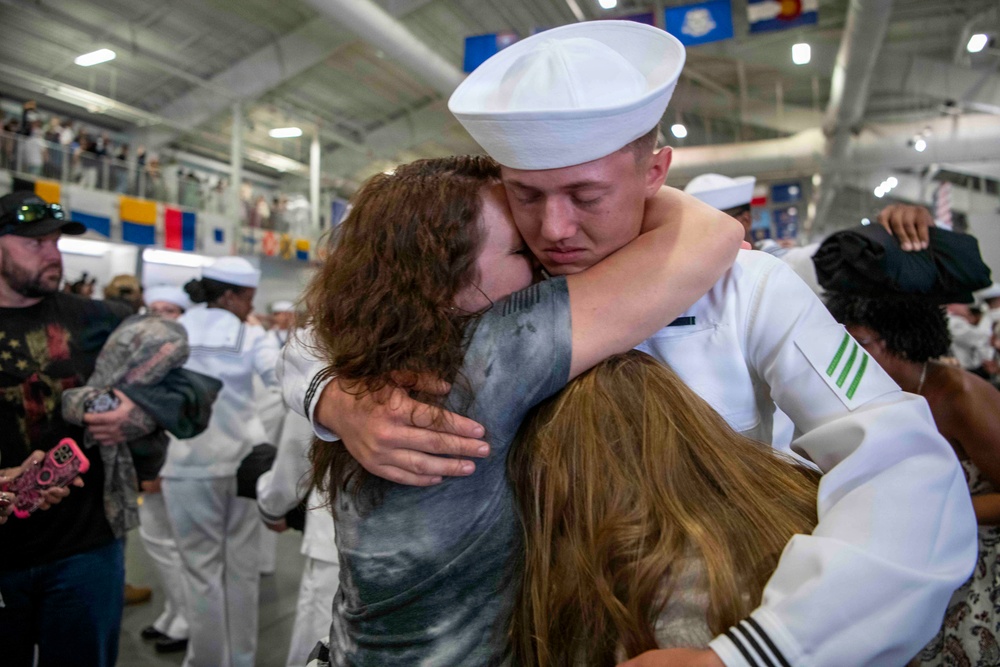 A U.S. Navy sailor in dress white uniform is embracing two people, likely family members, in an emotional reunion. The setting appears to be a military graduation or homecoming ceremony held in a large indoor facility. The moment captures a deeply emotional greeting with the sailor's eyes closed during the embrace.