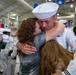 A U.S. Navy sailor in dress white uniform is embracing two people, likely family members, in an emotional reunion. The setting appears to be a military graduation or homecoming ceremony held in a large indoor facility. The moment captures a deeply emotional greeting with the sailor's eyes closed during the embrace.