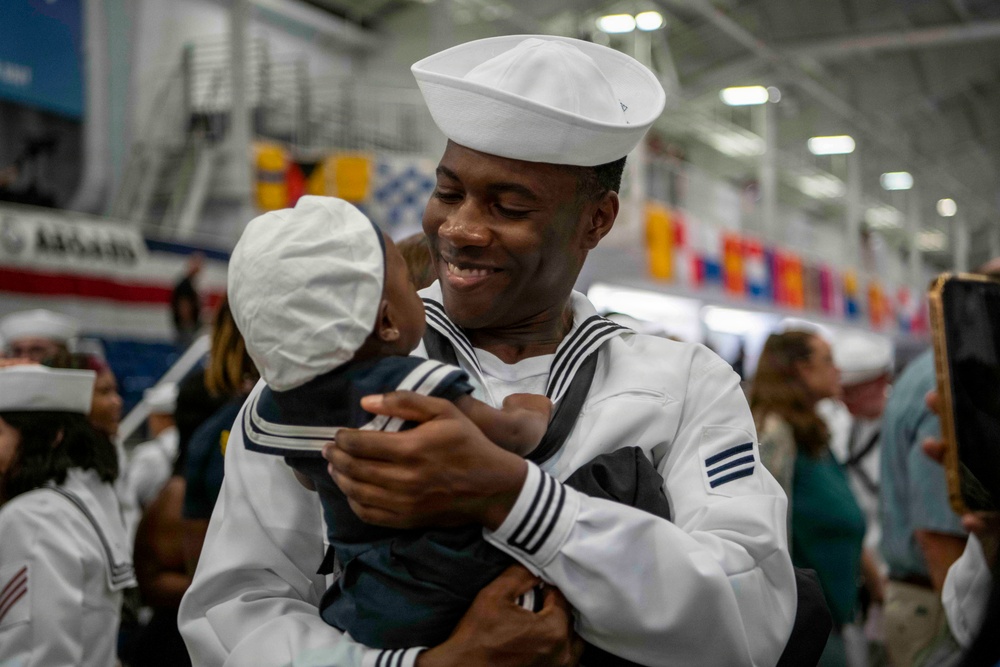 A U.S. Navy sailor in dress white uniform is holding and smiling at a young child dressed in a matching miniature sailor outfit. The setting appears to be an indoor military facility, likely during a graduation or homecoming ceremony. The joyful interaction suggests a family reunion or celebratory event.