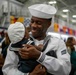 A U.S. Navy sailor in dress white uniform is holding and smiling at a young child dressed in a matching miniature sailor outfit. The setting appears to be an indoor military facility, likely during a graduation or homecoming ceremony. The joyful interaction suggests a family reunion or celebratory event.