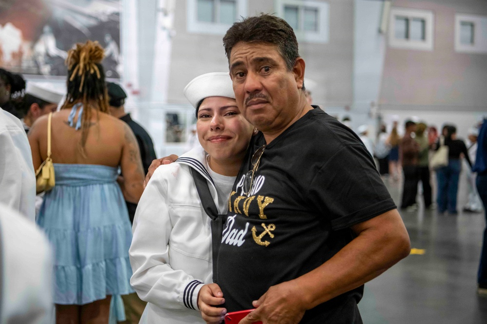 A female U.S. Navy recruit in dress whites embraces an older man wearing a black 'Proud Navy Dad' t-shirt, both showing emotional expressions consistent with a graduation or boot camp completion ceremony. The setting appears to be a large indoor military facility with other sailors and family members visible in the background. The moment captures a heartfelt reunion between a new sailor and her father.