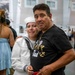 A female U.S. Navy recruit in dress whites embraces an older man wearing a black 'Proud Navy Dad' t-shirt, both showing emotional expressions consistent with a graduation or boot camp completion ceremony. The setting appears to be a large indoor military facility with other sailors and family members visible in the background. The moment captures a heartfelt reunion between a new sailor and her father.