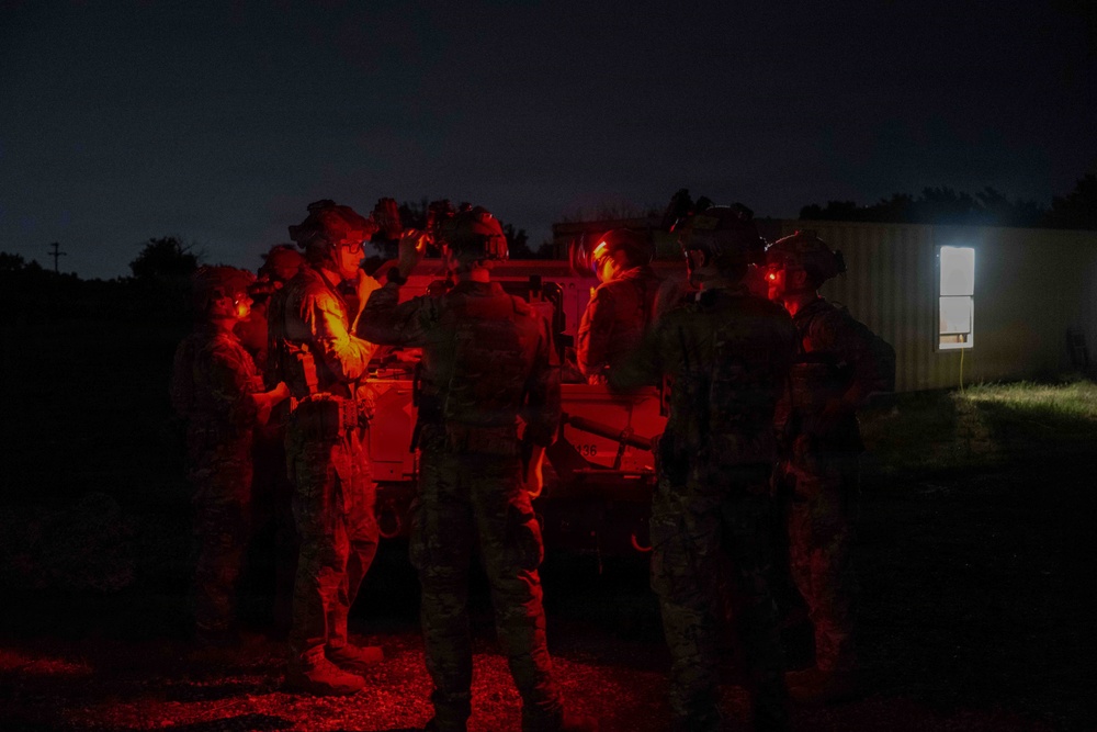 A group of military personnel in full combat gear are gathered around a vehicle at night, illuminated by red light. They appear to be conducting a briefing or mission planning session in a field environment. A lit structure is visible in the background to the right.