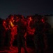 A group of military personnel in full combat gear are gathered around a vehicle at night, illuminated by red light. They appear to be conducting a briefing or mission planning session in a field environment. A lit structure is visible in the background to the right.