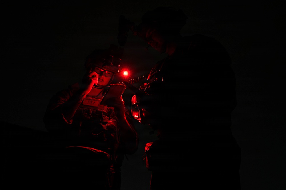 A nighttime military or tactical operation scene illuminated solely by red light sources. One individual wearing tactical gear, glasses, and a helmet-mounted device is visible, appearing to review documents or a map. A second figure is partially visible in the background, also in tactical equipment.