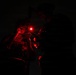 A nighttime military or tactical operation scene illuminated solely by red light sources. One individual wearing tactical gear, glasses, and a helmet-mounted device is visible, appearing to review documents or a map. A second figure is partially visible in the background, also in tactical equipment.