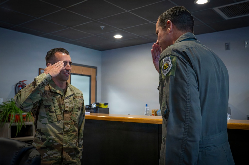 Two military personnel are exchanging salutes in what appears to be an indoor military facility or office. The individual on the left is wearing US Air Force OCP (Operational Camouflage Pattern) uniform, while the person on the right is wearing an olive green flight suit with a patch on the shoulder. Both are mid-salute, suggesting a formal military greeting or acknowledgment.