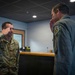 Two military personnel are exchanging salutes in what appears to be an indoor military facility or office. The individual on the left is wearing US Air Force OCP (Operational Camouflage Pattern) uniform, while the person on the right is wearing an olive green flight suit with a patch on the shoulder. Both are mid-salute, suggesting a formal military greeting or acknowledgment.