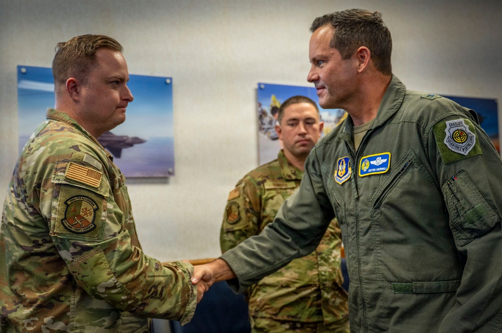 Two U.S. Air Force personnel are shaking hands in what appears to be an official recognition or greeting ceremony. The individual on the right is wearing a green flight suit with visible name tape reading 'Kevin Merrill' and multiple patches including wings and unit insignia. A third service member in camouflage uniform stands in the background observing the exchange.