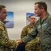Two U.S. Air Force personnel are shaking hands in what appears to be an official recognition or greeting ceremony. The individual on the right is wearing a green flight suit with visible name tape reading 'Kevin Merrill' and multiple patches including wings and unit insignia. A third service member in camouflage uniform stands in the background observing the exchange.