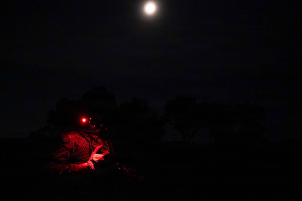 A nighttime image showing a person, likely a soldier or tactical operator, illuminated by a red light source. The individual appears to be in a prone or low position in an outdoor environment with tree silhouettes visible against a dark sky. A bright full moon or artificial light source is visible in the upper portion of the frame.