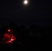 A nighttime image showing a person, likely a soldier or tactical operator, illuminated by a red light source. The individual appears to be in a prone or low position in an outdoor environment with tree silhouettes visible against a dark sky. A bright full moon or artificial light source is visible in the upper portion of the frame.