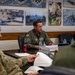 A military briefing or meeting is taking place in what appears to be a conference room at a U.S. Air Force installation. Name placards on the table identify Brig Gen Kevin Merrill, Commander Tenth Air Force, and Col Bradley Klemesrud. The room is decorated with historical photographs and unit insignia patches on the wall behind the central figure.