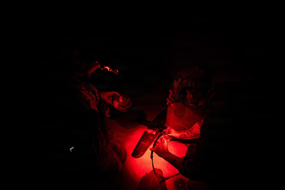 Two individuals are working together under red light illumination in an otherwise completely dark environment. They appear to be in military or tactical gear, examining or working with equipment on a surface between them. The red light source is positioned centrally, casting a deep crimson glow on their hands and the items they are handling.
