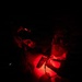 Two individuals are working together under red light illumination in an otherwise completely dark environment. They appear to be in military or tactical gear, examining or working with equipment on a surface between them. The red light source is positioned centrally, casting a deep crimson glow on their hands and the items they are handling.
