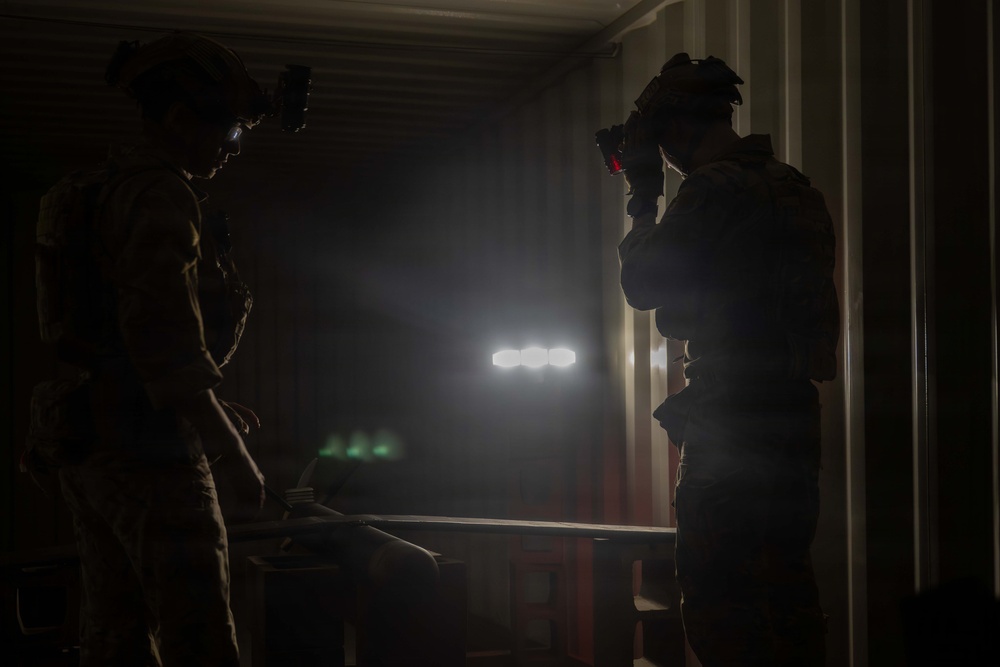Two military or tactical personnel are silhouetted in a dark, enclosed space that appears to be a shipping container or similar structure. One individual is raising an optical device (possibly binoculars or a camera) to their face, while both wear tactical helmets with mounted equipment. Bright artificial lights and hazy atmospheric conditions are visible in the background.
